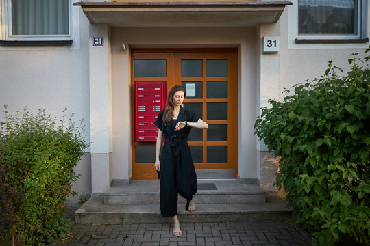 Woman in black jumpsuit checks her watch while standing on steps outside apartment building