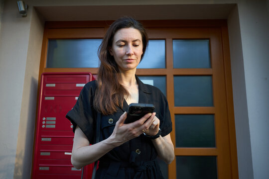 Woman in black outfit using smartphone outside apartment building with red mailboxes