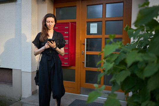Woman in black jumpsuit using smartphone while standing by apartment building entrance