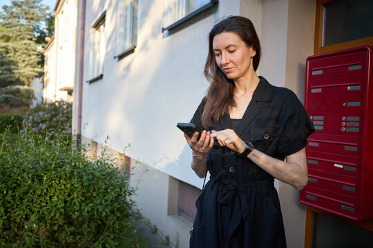 Woman using smartphone near red mailbox and building exterior in golden hour light