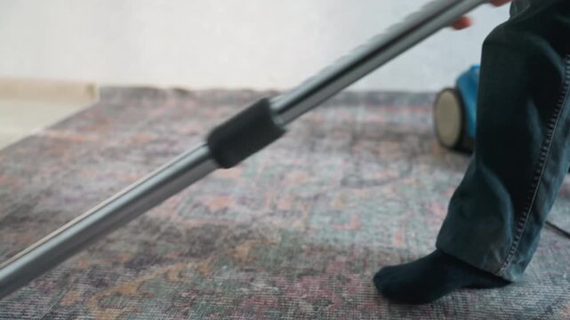 White man vacuuming patterned rug home, closeup of nozzle gliding across textured carpet, casual jeans and socked foot visible, natural light streaming, methodical strokes, quiet domestic cleaning
