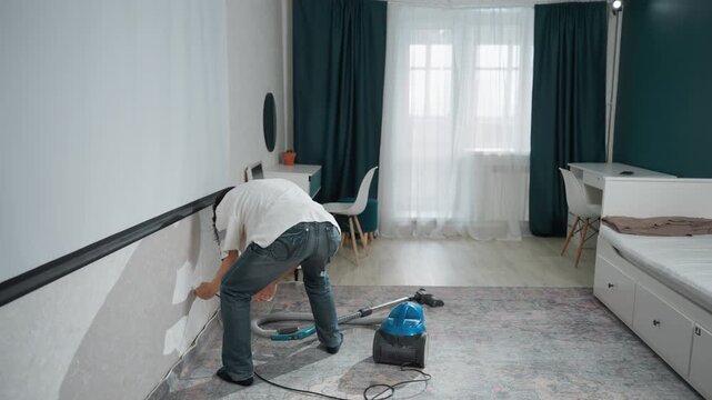 White man vacuuming along baseboard and carpet edge with nozzle attachment, focused homeowner leans to reach skirting and crevice lines, blue canister beside bed, methodical spot cleaning and stain