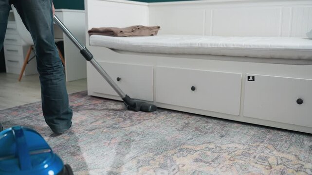 Vacuuming bedroom rug in home homeowner pushes blue canister vacuum across patterned carpet beside bed with storage drawers casual jeans and socks visible steady motion morning light tidy domestic