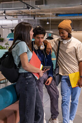 Diverse students in study lounge, viewing tablet, holding red and yellow folders with headphones © wavebreak3