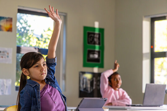 Asian female child wearing denim jacket raising right hand at classroom desk with tablet and laptop
