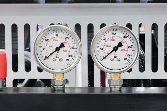 Two pressure gauges show 0 to 300 PSI readings. Round white dials have black numbers and red needles. Metallic bases sit on a black work surface. White slotted panel forms the background