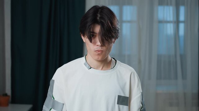 Asian man thoughtful pause in bedroom beside vacuum, downward gaze with moody lighting, introspective mood in minimal interior with desk and window, domestic routine interrupted, student vibe