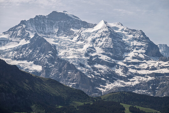 Dramatic Swiss Alpine peak with snowfields and rocky ridges under hazy summer sky, seen from the Lauterbrunnen Valley region.