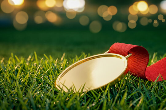 Gold medal blank face with red ribbon lying flat on green grass, football championship winner prize, warm golden bokeh stadium lights sport victory background