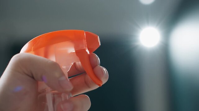 Spray bottle trigger releasing fine mist, closeup of hand gripping orange nozzle, steady press sends droplets into air, clear liquid and vapor catch cinematic light, demonstration of cleaning product