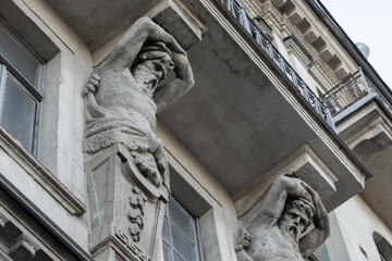 Obraz premium Weathered stone atlantes support a balcony on a historic building facade, showing classical muscular figures and intricate masonry details that convey age, craftsmanship and heritage of St.Peterburg