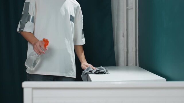 Young person wipes dresser with focus. Young individual attentively dusts white bedroom dresser surface. Teenage girl diligently using spray and cloth to tidy her bedroom furniture