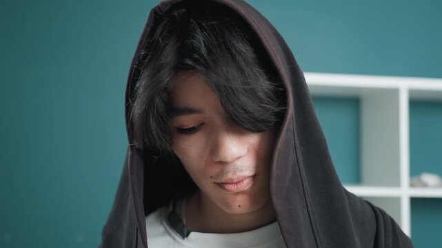 Asian man home face cleansing towel, young asian man wrapped in hooded towel at home applying facial cleanser and inspecting skin against teal shelf background quiet intimate bathroom moment showing