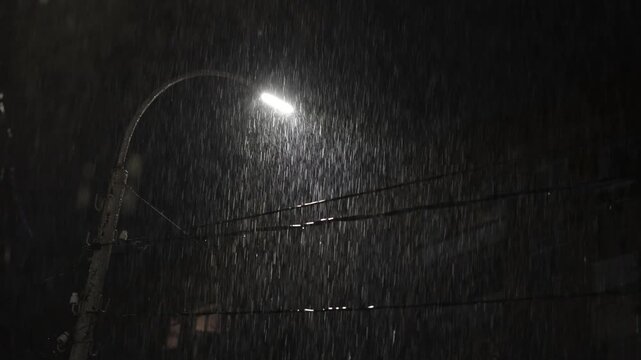 An atmospheric shot of a nighttime downpour on a city street. The bright beam of a streetlamp cuts through the darkness, vividly illuminating the falling raindrops and creating dramatic contrasts. 