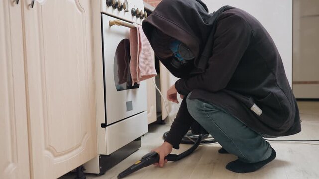 Professional cleaner vacuuming under oven, hooded person in jeans kneels to reach debris beneath appliance, crevice nozzle, targeted suction, realistic home maintenance, bright kitchen