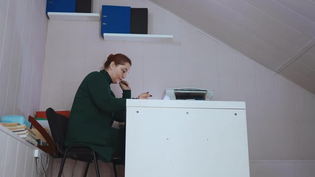 Home office paramedic making emergency call woman in green uniform seated at attic desk with printer and stacked files, dialing phone while organizing paperwork and laptop, focused and concerned