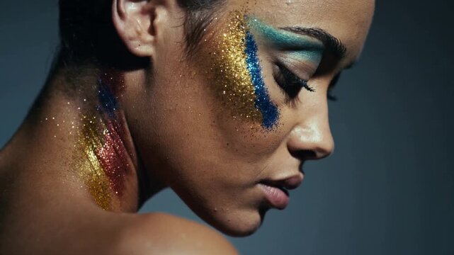Close-up of a person's face and neck. Gold glitter decorates the cheek. Blue and red glitter lines the neck. Dark gray background contrasts the colors. The person shows a serious expression