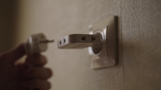 Close-up hand of unrecognizable person plugging white power adapter into wall socket, device inserted to connect electricity. Concept of energy consumption, power, connection in home.
