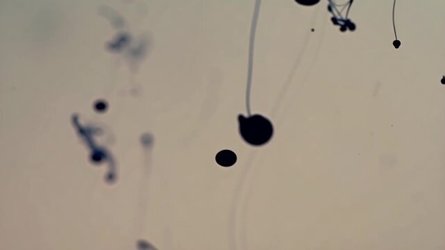 Dark ink drops falling into water, dispersing and dissolving slowly to create abstract fluid shapes and smoke-like patterns on a light background.