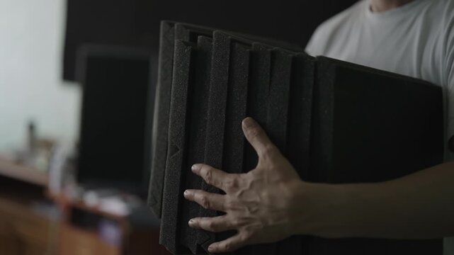 Focused man in white t shirt holding and inspecting stack of black sound absorbing foam panels, preparing for acoustic treatment of music studio or recording booth. Shooting in slow motion.