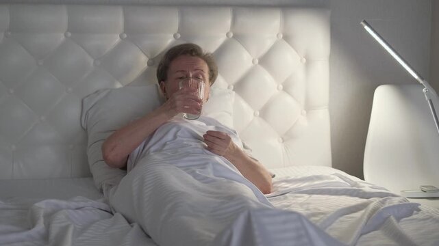 An elderly woman takes a pill before going to bed while lying in bed