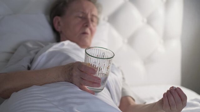 An old woman holds a pill and a glass of water in her hands while lying in bed, close-up