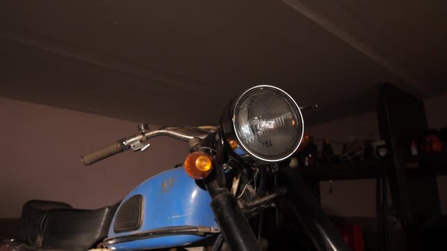 Close up view of a classic vintage motorcycle's headlight, front forks, and handlebars, showing its worn blue fuel tank and an overall retro style in a dimly lit garage