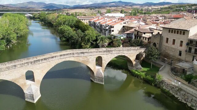 A drone view of the ancient Puente la Reina stone bridge crossing Arga River, with trees, buildings, and distant mountains under a bright sky in Spain
