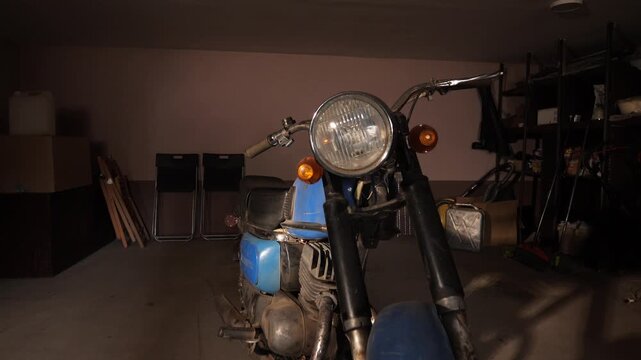 Vintage blue motorcycle with a round headlight parked in a dim garage, representing themes of nostalgia, mechanics, and classic transportation, sitting among various stored items and tools