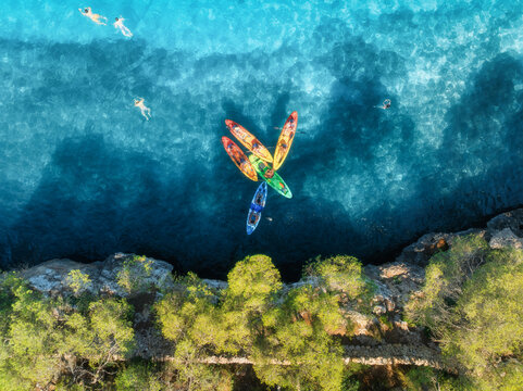 Aerial drone view of colorful kayaks in crystal clear turquoise sea near rocky coast and green trees. Summer outdoor adventure in Mallorca, Balearic Islands, Spain. Top down view of canoes. Vacation	