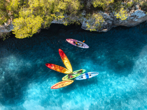 Aerial drone view of colorful kayaks in crystal clear turquoise sea near rocky coast and green trees. Summer outdoor adventure in Mallorca, Balearic Islands, Spain. Top down view of canoes. Vacation	