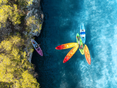 Aerial drone view of colorful kayaks in crystal clear turquoise sea near rocky coast and green trees. Summer outdoor adventure in Mallorca, Balearic Islands, Spain. Top down view of canoes. Vacation	