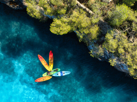 Aerial drone view of colorful kayaks in crystal clear turquoise sea near rocky coast and green trees. Summer outdoor adventure in Mallorca, Balearic Islands, Spain. Top down view of canoes. Vacation	