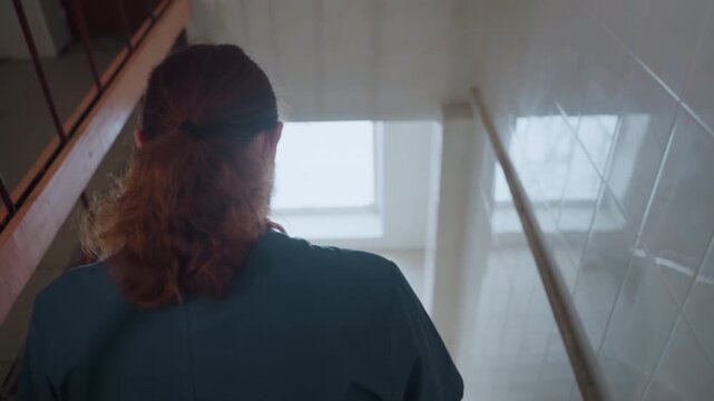 White paramedic with red ponytail at stairhead, rear silhouette framed by skylight, pausing to assess scene and adjust stethoscope, preparing for descent while checking equipment, reflective