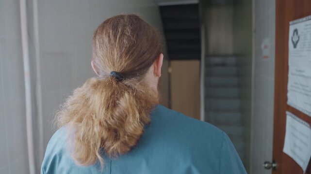 Paramedic walking down hospital corridor, scrubs. Tiled walls, numbered doors, soft daylight through window, ponytail tied at nape, quiet tension of night shift, clinical posters on walls, stairwell