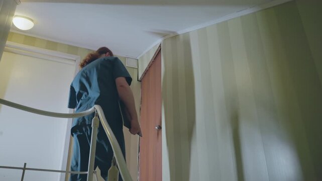 Home nurse climbs upstairs railing and approaches bedroom door, patient support scenario with careful ascent, dim landing light and bedside frame visible, attentive caregiver motion