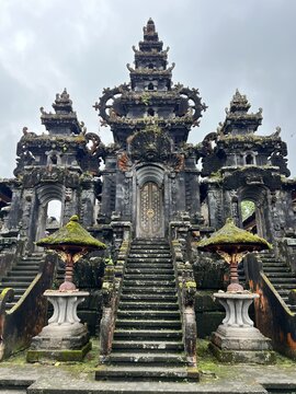 Ornate stone staircase and elaborately carved gateway of Pura Besakih, the Mother Temple of Bali, Indonesia, covered in moss and tropical vegetation under an overcast sky
