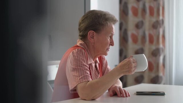 An elderly sad pensive woman in the kitchen drinking tea