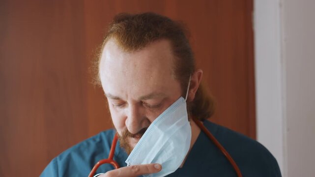 Home responder removing mask, postshift fatigue evident white male peels ear loops while stethoscope rests on neck, reflective pause in doorway, weary eyes and quiet decompression after house call,