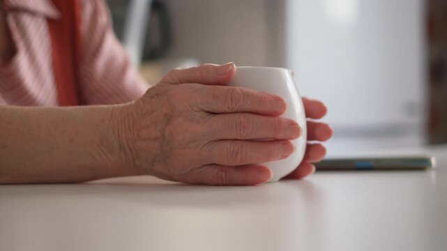 An elderly woman warms her hands on a hot cup of tea, close-up