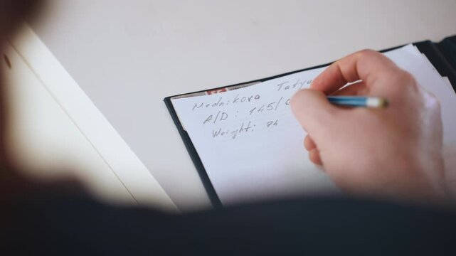 Paramedic writing patient notes on clipboard, closeup of hand holding pen, document showing scribbled observations, inside quiet home during house visit, blurred shoulder foreground, clinical
