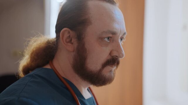 Concerned white paramedic in house, focused bedside assessment with stethoscope, beard and ponytail, soft window light, clinical uniform, quiet tension and empathy during home visit
