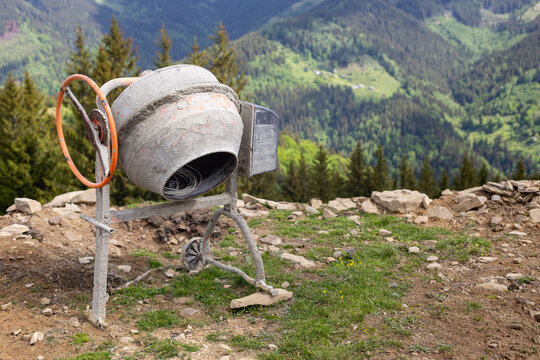 Concrete mixer on mountain construction site. Portable concrete mixer stands on rocky ground at a mountain construction site overlooking a green forested valley.