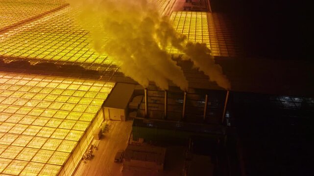 Vast illuminated greenhouse complex at night, glowing with powerful grow lights as thick steam rises from industrial chimneys, seen in aerial views of indoor agriculture