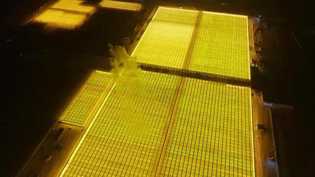 Drone night view of expansive illuminated greenhouse complex with glowing golden panels forming geometric grids against surrounding darkness, showcasing advanced horticulture