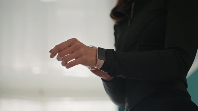 Woman adjusting smartwatch before home workout, standing by bright window, black activewear, focused posture, checking timer and notifications, preparing for highintensity session or virtual class