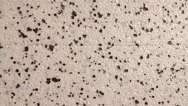 Fotografia przedstawia zbliżenie styropian o jasnobeżowej, chropowatej strukturze. Na powierzchni widoczne są liczne, drobne otworki oraz nieregularne, ciemne nakropienia i punkty.