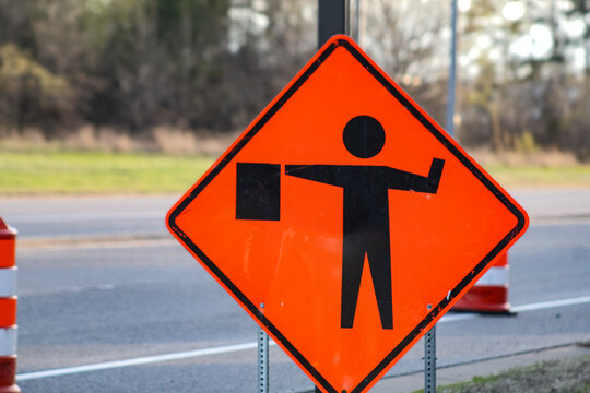 Orange diamond-shaped road construction flagger symbol warning sign on the side of a street with blurred background