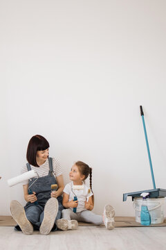 A mother and her young daughter sit on the floor, holding paint rollers and brushes, ready to redecorate a room