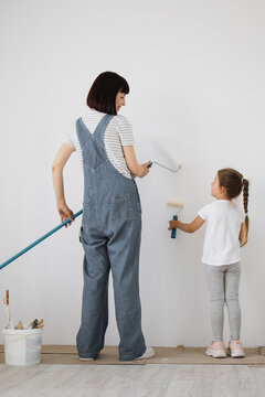 A woman and a young girl in overalls are painting a white wall with rollers, engaged in a home improvement project together